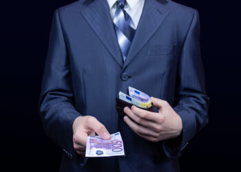 A person in a dark suit and striped tie holds a wallet with several euro banknotes in one hand and offers a 500-euro note—resembling the thrill of UK Casino Winnings—against a dark background. | FintechZoom