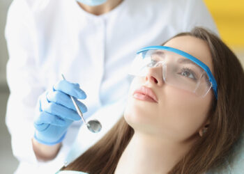 A woman wearing protective glasses lies in a dental chair, looking up, while an endodontist in gloves and a white coat holds a dental mirror near her mouth. | FintechZoom