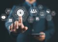 A person in a suit touches a virtual “PAYROLL” icon on a transparent screen displaying various business, finance, and human resources icons. The person is holding a tablet in the other hand. | FintechZoom