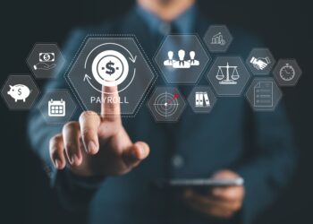 A person in a suit touches a virtual “PAYROLL” icon on a transparent screen displaying various business, finance, and human resources icons. The person is holding a tablet in the other hand. | FintechZoom
