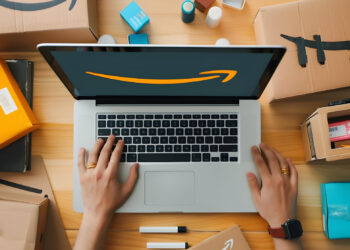 A person types on a laptop displaying the Amazon logo, surrounded by Amazon packages, boxes, and small parcels on a wooden table. | FintechZoom