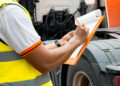 A person wearing a reflective safety vest writes on a clipboard while standing next to a large truck, possibly performing an inspection or checklist as THC testing affects commercial drivers. | FintechZoom