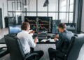 Two men in business attire sit at a desk in a modern office, analyzing financial charts and stock market data on multiple monitors. They appear to be discussing market trends and effective risk management strategies while taking notes. | FintechZoom