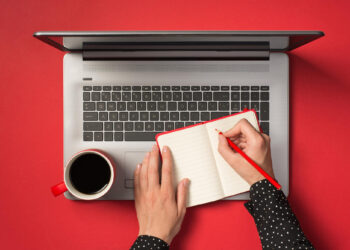 A person writes in a notebook with a red pencil while resting it on a laptop keyboard; a cup of coffee sits nearby on a bright red desk. | FintechZoom