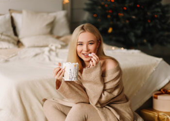A smiling woman in cozy beige loungewear sits by a bed, holding a festive mug and marshmallow. A decorated Christmas tree and wrapped gifts are in the background, creating a warm, holiday atmosphere. | FintechZoom