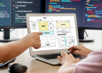 Two people work together at a desk, looking at a laptop displaying a website wireframe. One person points at the screen while the other types. Computer monitors with code are visible in the background. | FintechZoom