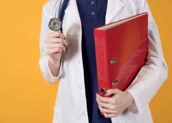 A person in a white lab coat holding a stethoscope in one hand and a large red binder in the other, standing against a bright yellow background. | FintechZoom