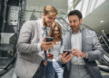 Three businesspeople in formal attire stand in front of escalators, smiling and engaged as they discuss something on their smartphones—possibly the convenience of using virtual cards—in a modern office building or mall. | FintechZoom