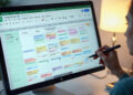 A person uses a stylus to organize a colorful, detailed schedule on a spreadsheet displayed on a large computer monitor. A lamp glows softly in the background, creating a focused work atmosphere. | FintechZoom