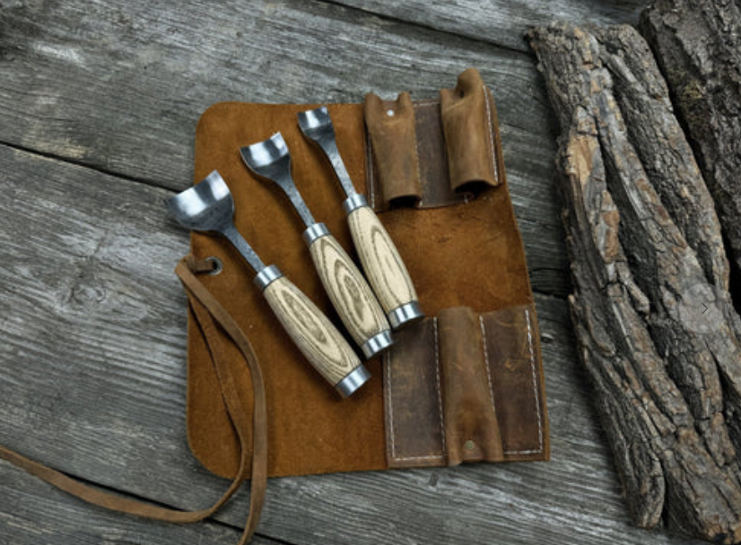 Three wood carving tools with light wooden handles and metal scoops are displayed on an open brown leather tool roll, resting on a weathered wooden surface next to a piece of rough tree bark. | FintechZoom