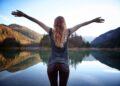A person with long wavy hair stands at the edge of a calm lake, arms outstretched, facing distant forested mountains reflected in the water under a clear sky. | FintechZoom