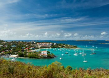 A scenic bay in Antigua and Barbuda with turquoise water, white sailboats anchored near a coastal town of colorful buildings, lush greenery, and a blue sky with scattered clouds. | FintechZoom