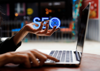 A person types on a laptop while holding a glowing “SEO” text with a gear icon above their hand, symbolizing Fintech SEO and digital technology in search engine optimization. | FintechZoom