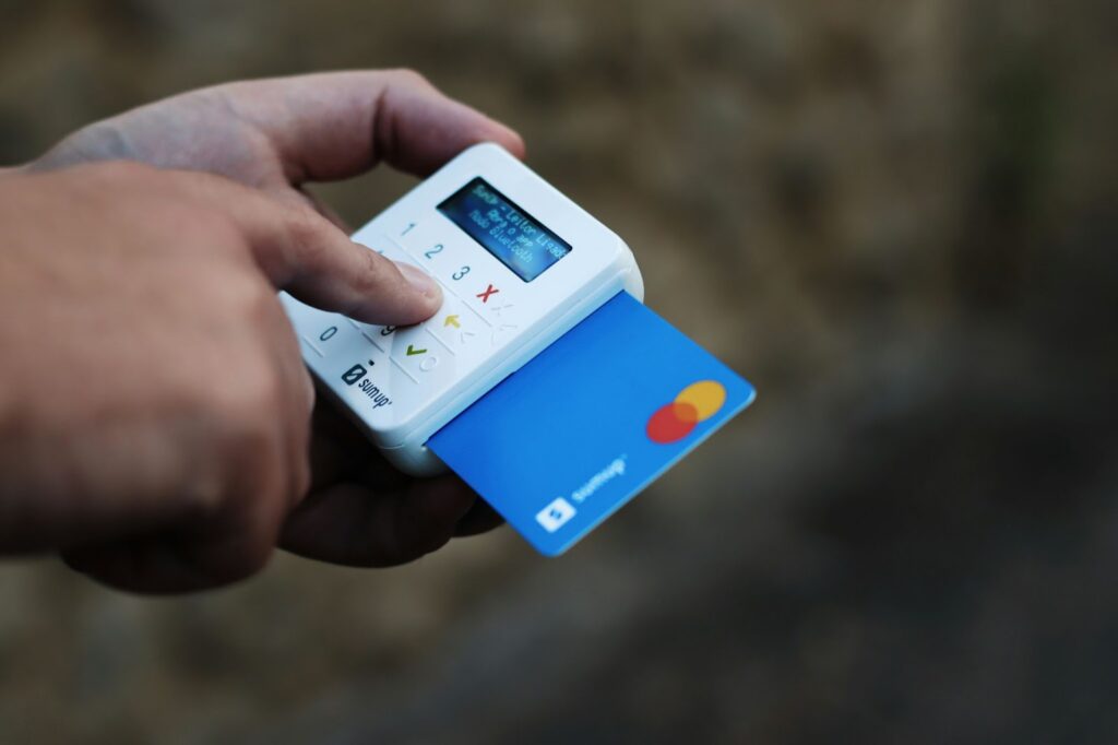 A hand holds a small white card reader with a blue Corporate Card partially inserted. The device displays text on a small screen, while the background remains blurred. | FintechZoom