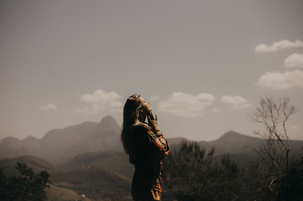 A woman stands outdoors with her eyes closed and head tilted upward in peaceful acceptance. Distant mountains rise under a hazy sky with scattered clouds, while dry branches softly frame the serene scene. | FintechZoom