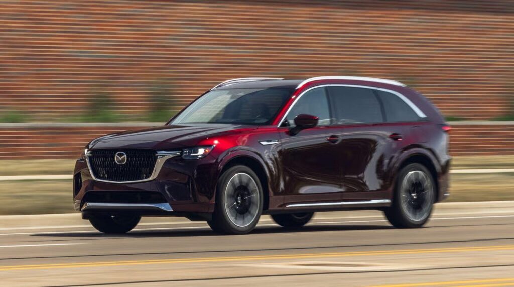 Mazda CX-90 dark red SUV speeds down the road, with a blurred brick wall background highlighting its motion and power. | FintechZoom