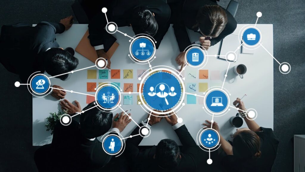 A group of people in business attire gather around a table with papers and sticky notes, while digital icons representing teamwork, planning, HR software, and technology are superimposed over the scene. | FintechZoom