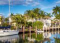 A white sailboat docked by waterfront homes with palm trees reflected in the calm water under a blue sky.
