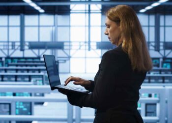Woman in a suit using a laptop in a modern server room with rows of computer servers in the background.
