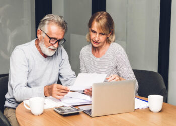 An older man and woman sit at a table with papers, a laptop, a calculator, and two coffee mugs, appearing focused as they review documents together. | FintechZoom