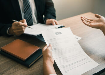Two people sit at a desk during a job interview; one person holds a resume while the other, dressed in a suit, takes notes on a clipboard. A notebook and folder are on the desk. | FintechZoom