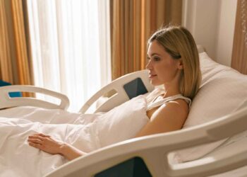 A woman with blonde hair lies in a hospital bed, propped up with pillows, looking thoughtful by a sunny window.