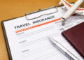 A travel insurance form on a clipboard with blank fields, a silver pen, a maroon passport, a small toy airplane, and a boarding pass on a wooden surface. | FintechZoom