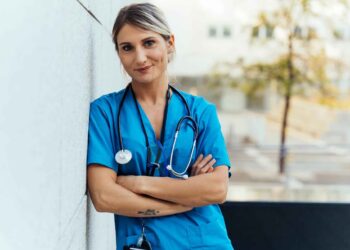 A healthcare professional in blue scrubs with a stethoscope around their neck stands outdoors, leaning against a wall with arms crossed, smiling confidently at the camera. | FintechZoom