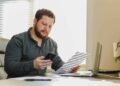 Man sitting at desk, looking at phone and holding a paper, with a laptop open in front of him.
