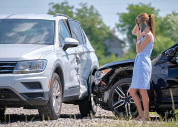 A woman in a light blue dress stands by two damaged cars after an accident, holding a phone to her ear. One car is white with a dented side; the other is black with a damaged front. | FintechZoom
