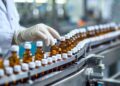 A worker in gloves inspects brown glass bottles with white caps on a conveyor belt in a pharmaceutical lab.