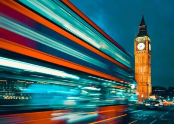 A red double-decker bus speeds past Big Ben in London at night, with bright city lights and motion blur.