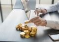 Person working on a laptop with gold bars and gold coins on the desk next to a smartphone.