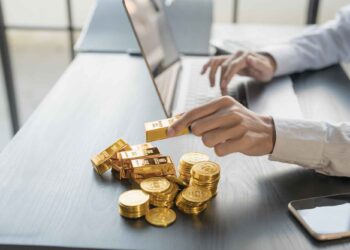 Person working on a laptop with gold bars and gold coins on the desk next to a smartphone.