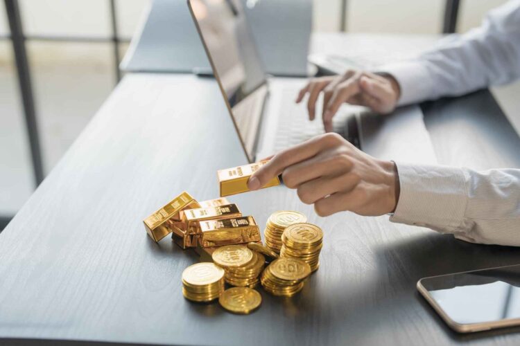 Person working on a laptop with gold bars and gold coins on the desk next to a smartphone.