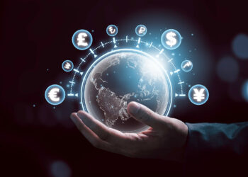 A hand holds a glowing digital globe showing continents, surrounded by floating currency symbols such as dollar, euro, pound, yen, and bitcoin, representing global finance and digital economy. | FintechZoom