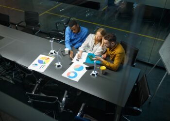 Three people sit at a conference table in an office, reviewing charts and graphs on paper. They are focused on their discussion, with coffee cups and documents spread out in front of them. | FintechZoom