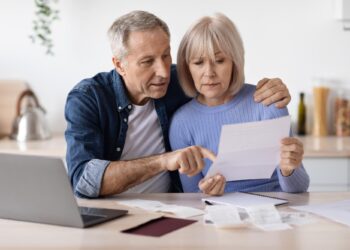 Older couple sitting at a table reviewing documents with a laptop, looking concerned.