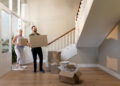 A man and a woman carry cardboard boxes into a bright, empty room with wooden floors and a staircase, suggesting they are moving into a new home. Other unpacked boxes and packing materials are on the floor. | FintechZoom