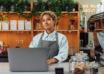 A barista wearing a gray apron stands behind a counter with a laptop in a cozy café with plants and jars.