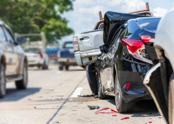 A black car and a white pickup truck are damaged from a rear-end collision on a busy road in Iowa, with debris scattered on the ground—typical of Car Accidents in Iowa—and other vehicles visible in the background. | FintechZoom
