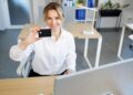 A woman in a white shirt sits at a desk in an office, smiling and holding a black card up to the camera. A computer monitor and office supplies are visible on the desk. | FintechZoom
