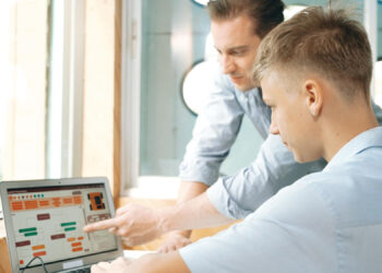 Two men are sitting at a desk by a window, looking at a laptop screen with colorful charts and diagrams. One man is pointing at the screen while the other listens attentively. | FintechZoom