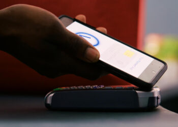 A person holds a smartphone over a card reader, using a contactless payment app to make a purchase. The card reader sits on a counter, and the phone screen displays a payment interface. | FintechZoom