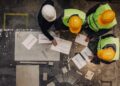 Overhead view of construction workers in safety gear reviewing blueprints on a table at a worksite.