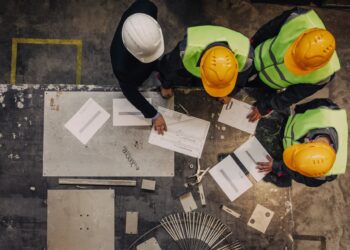 Overhead view of construction workers in safety gear reviewing blueprints on a table at a worksite.