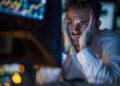 A worried man in a white shirt stares at computer screens displaying financial charts and data.