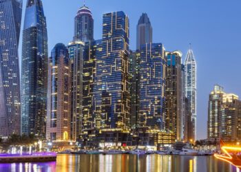 Dubai Marina skyline at dusk, featuring modern high-rise buildings lit with decorative lights, reflections shimmering on the water, and docked boats along the waterfront. | FintechZoom