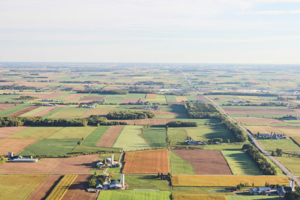 Aerial view of rural property reveals a patchwork of green, brown, and yellow farmland divided by roads and tree lines under a clear sky, with scattered farm buildings and open fields stretching into the distance. | FintechZoom