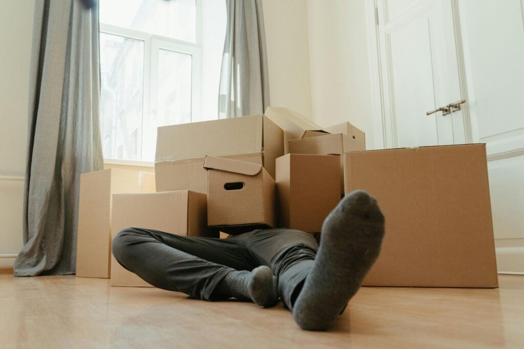 A person lies on a wooden floor surrounded by several cardboard moving boxes, taking a break during a long-distance move in a bright room with large windows and light-colored curtains. | FintechZoom
